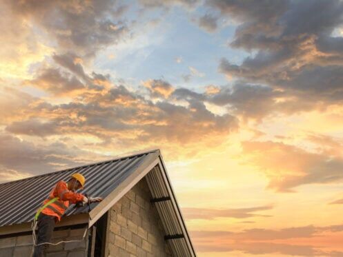 how to install metal roofing over shingles-roofer in safety gear standing on ladder installing metal roofing on a house