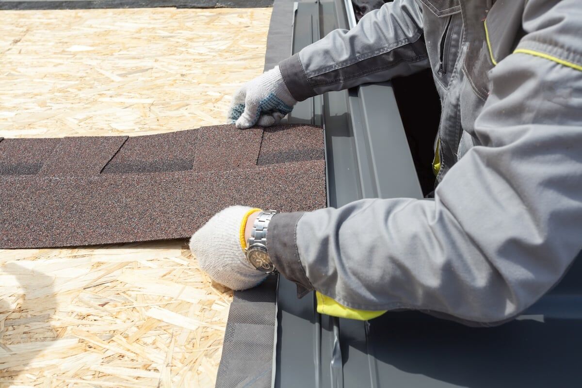 Cropped view of professional roofer worker lays asphalt sheet on the rooftop, holding roof tile in hands