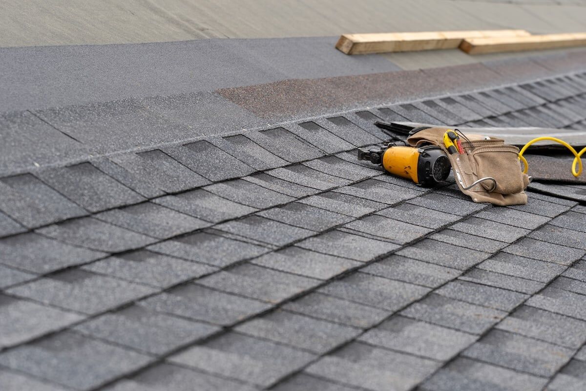 Photo of toolbelt with instrument and nail gun lying on asphalt or bitumen shingle on top of the new roof under construction residential house or building