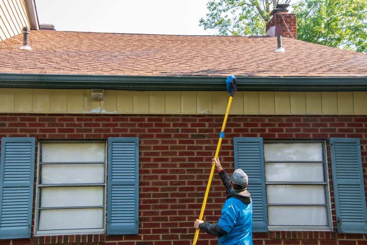 Young man cleaning the soffit of a house with a brush on a long pole. 